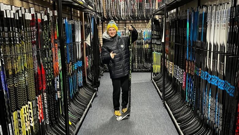 A smiling teenage boy holds up a hockey stick in one hand, pointing with the other, in the middle of a store aisle full of hockey sticks. 