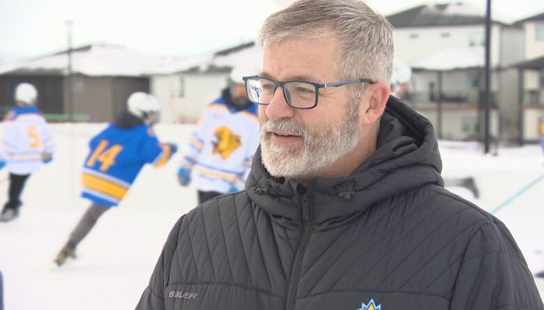 An older man with glasses and a winter jacket looks stands in front of an outdoor ice rink where hockey players are playing in the background.