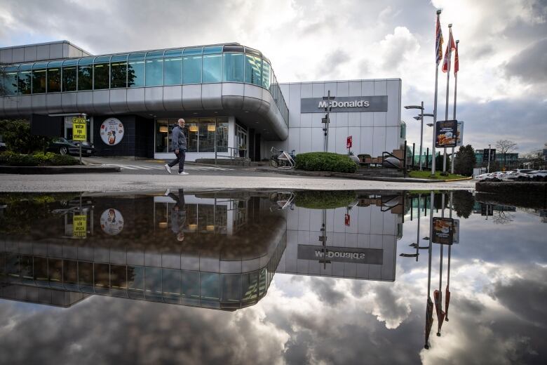 A man walks past a large puddle, in which the reflection of a McDonald's restaurant is visible with clouds in the sky and some flags off to the right.