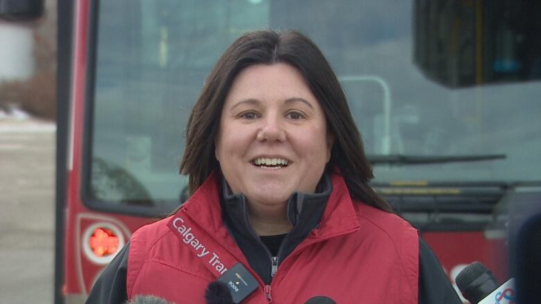 A woman with shoulder-length dark hair smiles while talking to reporters. She is wearing a red Calgary Transit vest over a black jacket. 