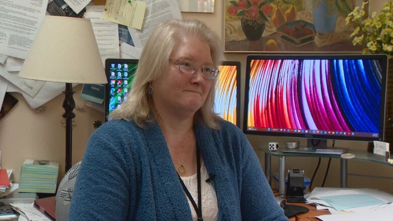 A woman is seen from the chest up sitting in front of her desk and monitors. She's wearing a blue cardigan and white shirt. 