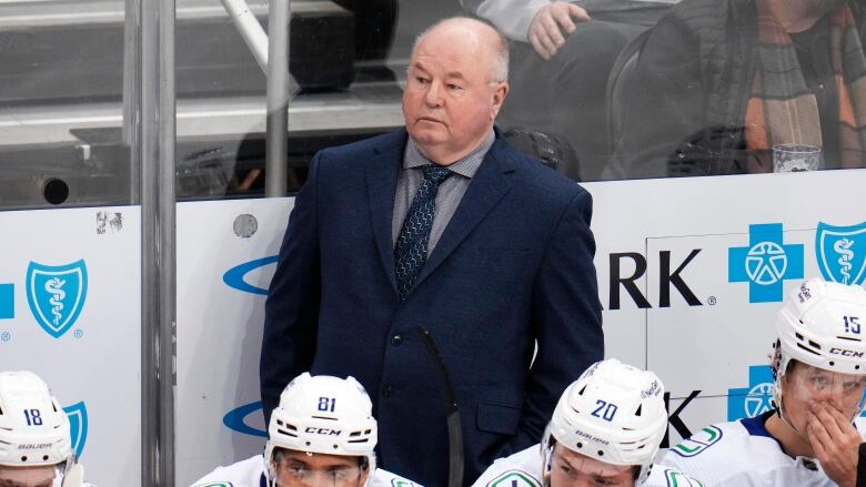 A coach stands behind the bench, with four Canucks players sitting in front of him.