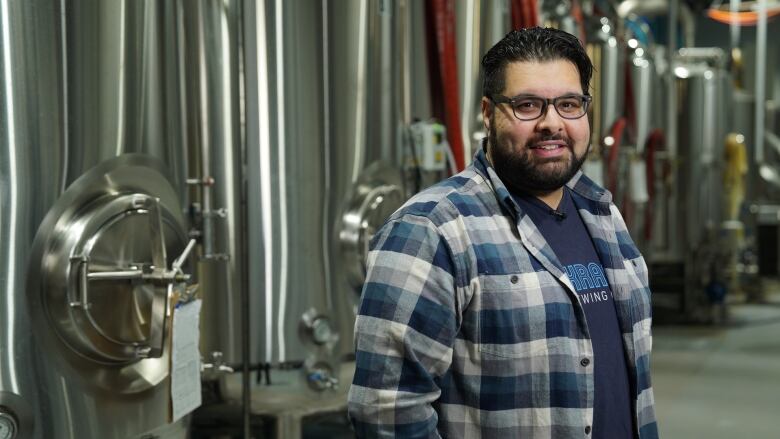 A man with short dark hair and beard is wearing glasses and a blue and grey plaid shirt. He stands smiling in front of a long row of cylindrical brewing machinery.