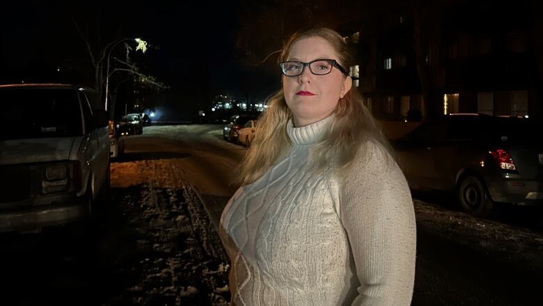 A blonde woman stands in an empty street at night, looking into the camera, wearing glasses and a white knit turtleneck sweater.