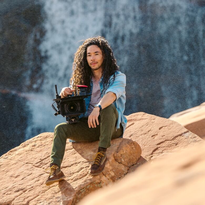 A man holding a large camera sits on a big rock in front of a waterfall