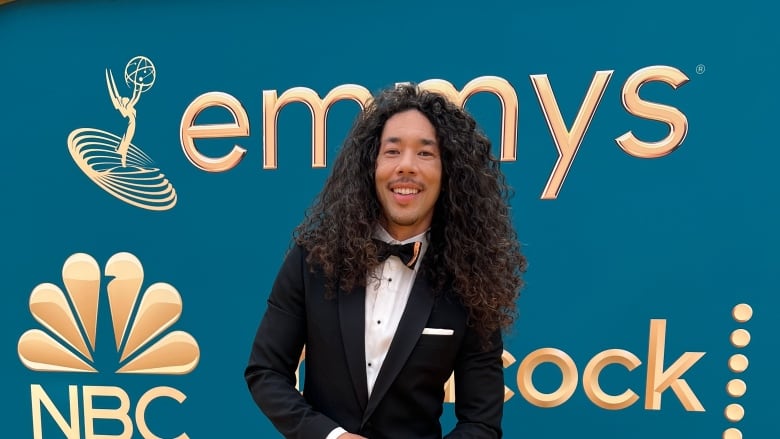 A man with long curly black hair wearing a suit smiles in front of a sign that says Emmys.