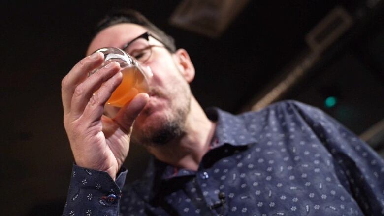 A man wearing a navy button-down and glasses sips an amber-coloured drink from a glass tumbler.