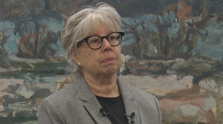 A woman in a checked blazer stands in front of a painting. she has white hair 