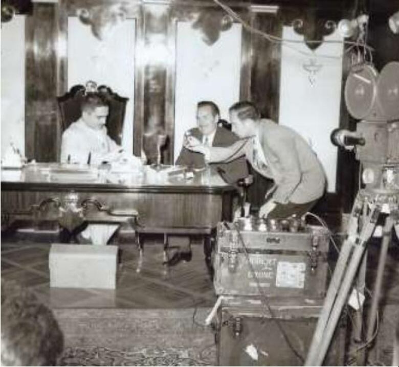 A black-and-white picture of a cameraman reaching a mic across a table, with a seated person in general's uniform and a a smiling man.