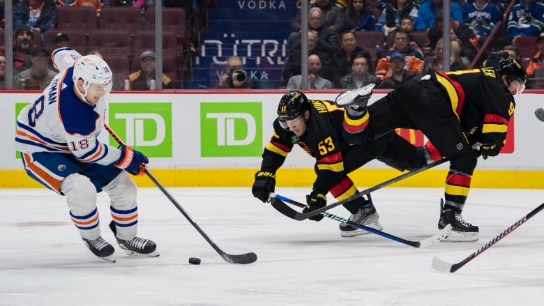 An Edmonton Oilers player conducts the puck in the left side of the picture as two Vancouver Canucks players appear to be losing their balance as they head to opposite ways in the right side of the pictures. 