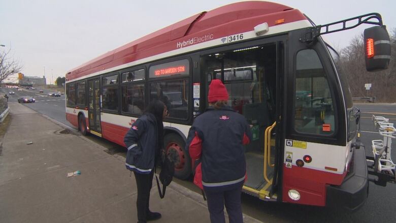 Two TTC operators in uniform stand near the open doors of a TTC bus.