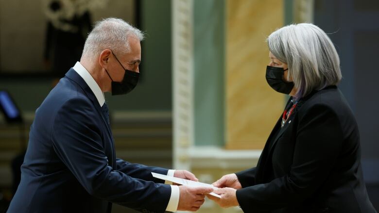 A man, wearing a mask, hands a piece of paper to a woman, also masked.