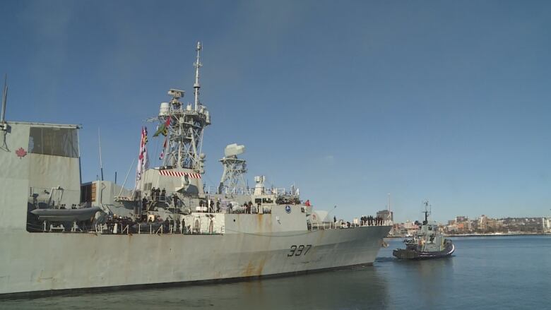 A frigate is being towed out of harbour by a tugboat.