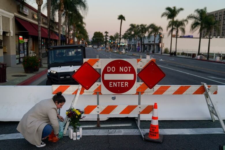 A woman kneels before flowers that are laid in front of a road block barrier.