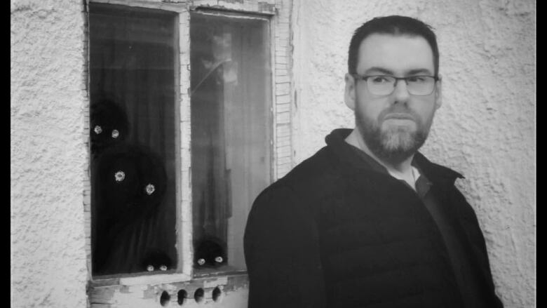 A black and white image of a man next to a window with some drawings of shadows behind the window.