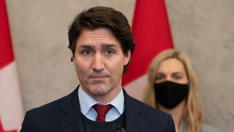 A man in a suit and tie stands at a podium while a woman stands behind him wearing a mask. 