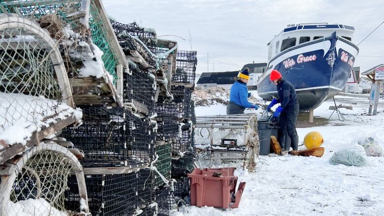 Workers gather lobster traps and rope on shore with a large fishing boat in the background.