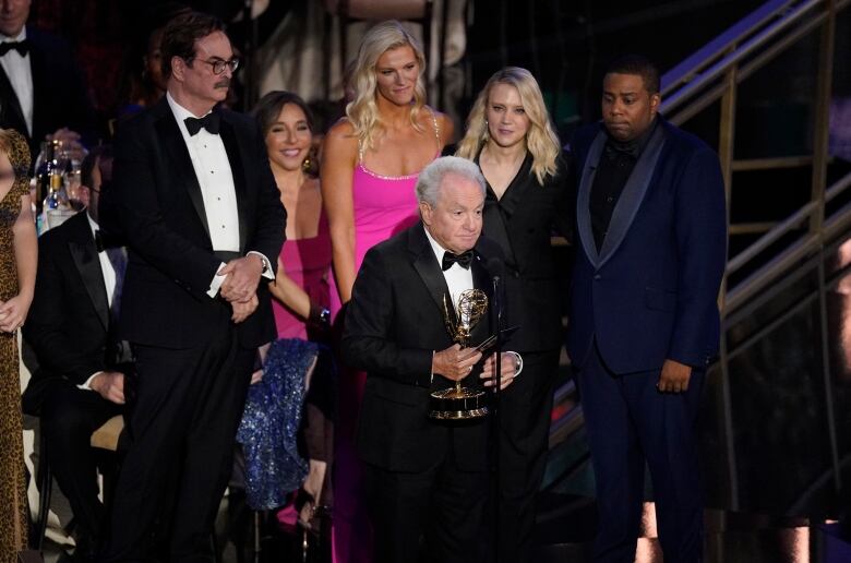 A man holding an award gives a speech into a microphone on a stage while a group of other people stand behind him.