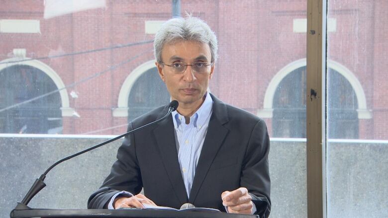 A white-haired man in a dark suit with an open-collared white shirt stands at a podium in front of a window.