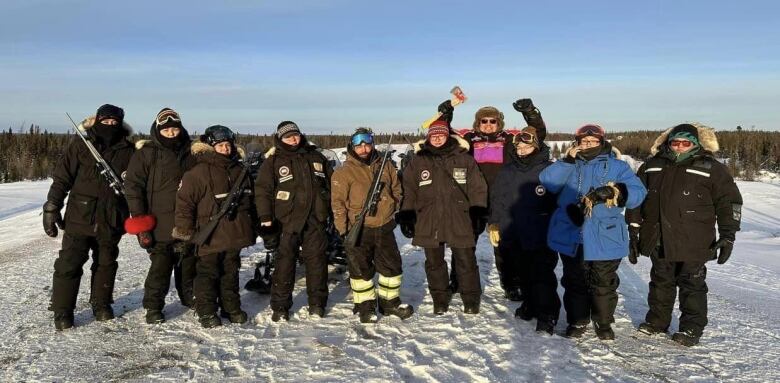 A large group in snowmobile gear and with rifles over their shoulders celebrates a successful hunt outside on a wintery day in northern Quebec.