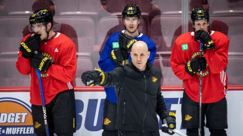 A man dressed in black, identified as the new coach, holds a hockey stick pointing toward the ice while three players stand behind him during hockey practice.