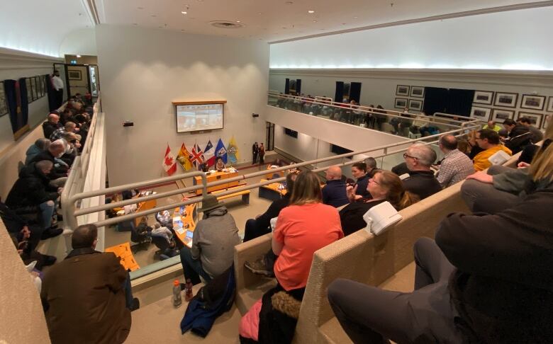 A group of people gather sit overlooking a city hall council chamber.