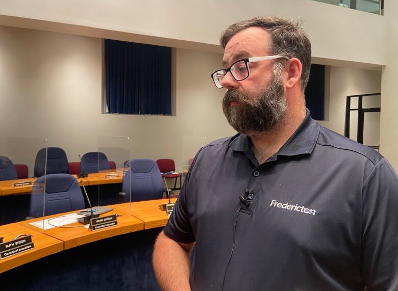 A bearded man wearing a polo shirt with the word Fredericton on it stands in a council chamber. 