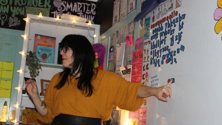 woman with dark hair and glasses pointing to a white board with letters on it