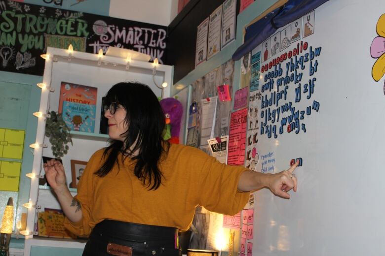 woman with dark hair and glasses pointing to a white board with letters on it