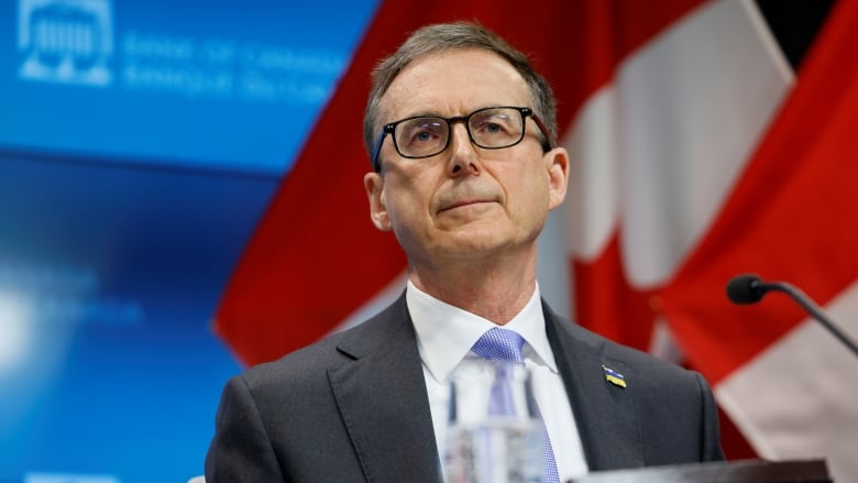 A man sits in front of a microphone with a Canadian flag and 'Bank of Canada' visible on the background behind him.