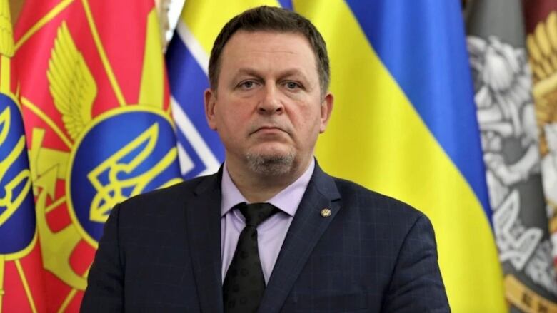 A man in a suit looks grim-faced standing in front of several different flags of blue, yellow, red and grey.