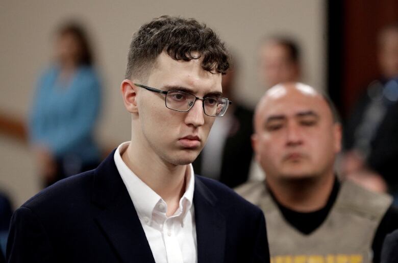 A young man in glasses wearing a suit jacket and collared shirt is shown indoors.