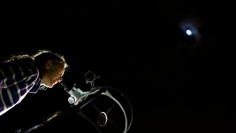 The photo shows a girl wearing a striped shirt looking through a telescope a the moon against a jet black sky. 