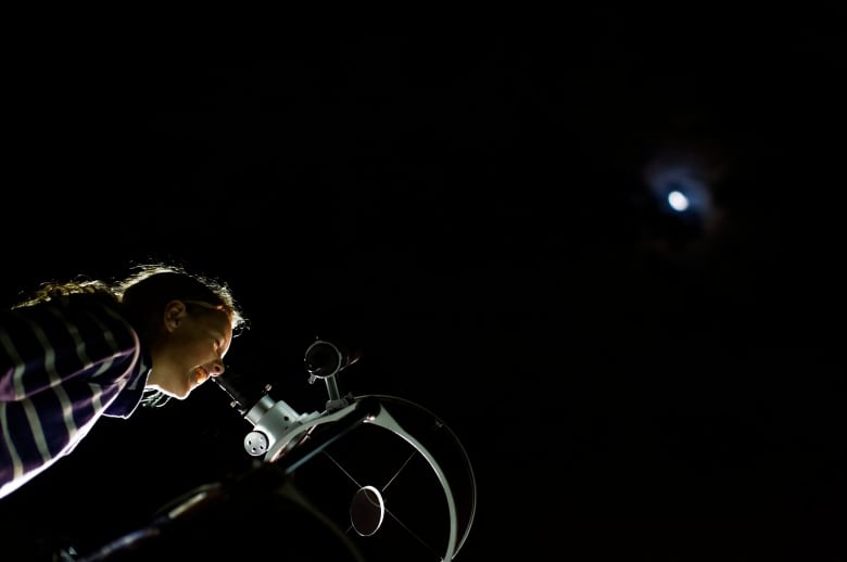 The photo shows a girl wearing a striped shirt looking through a telescope a the moon against a jet black sky. 