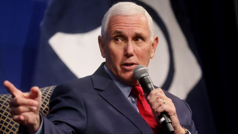 A man in a blue suit and red tie is seen speaking into a microphone.