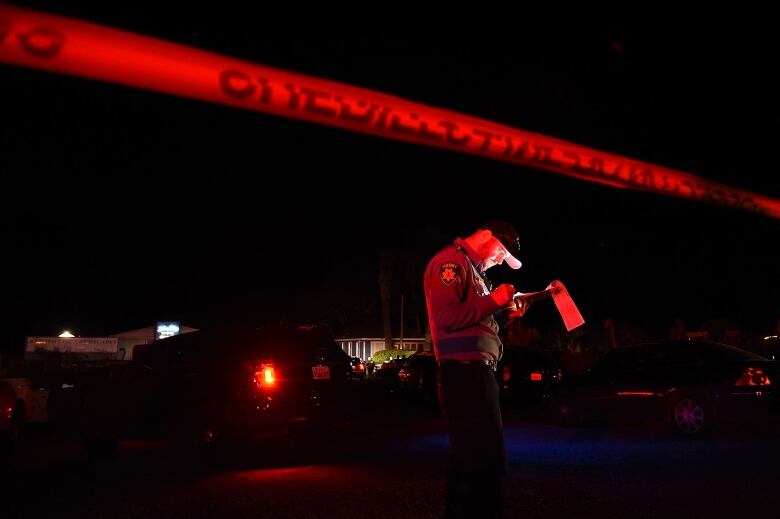 A man who appears to be writing on a clipboard standings behind a police line in the dark. The nighttime lighting has him looking red.