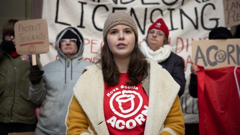 A woman with brown hair in a fuzzy hat and coat stands in front of a group of people holding signs. The signs read 'protect tenants' and call for landlord licensing.