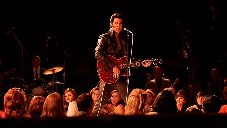 A man with dark hair sings and plays the guitar at a microphone on a stage, while a group of young people look up.