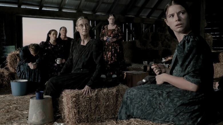 A group of women sit on bales of hay in a barn.