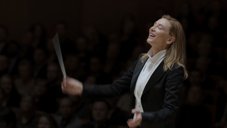 A woman with long hair in a black suit and white shirt holds a conductor's baton.