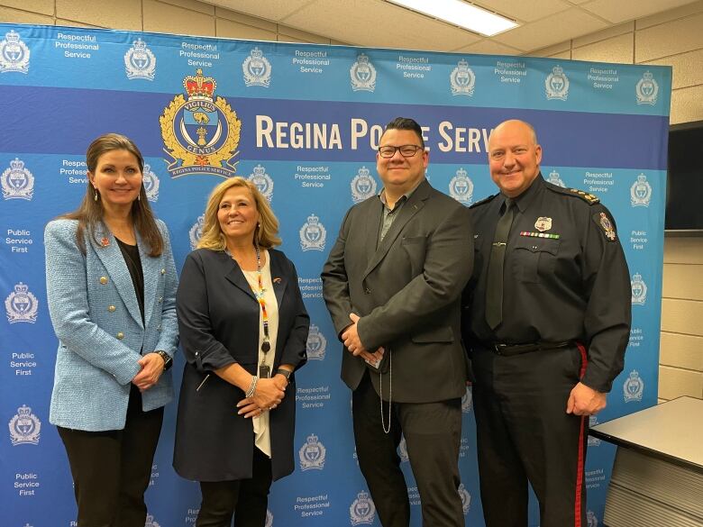 Four people -- two women on the left, two men on the right -- stand side-by-side smiling toward the camera. They're standing in front a blue poster that reads, 'Regina Police Service,'  and features the service's logo.