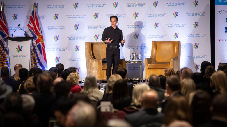 A man speaks to a crowd in front of two armchairs and a small table with a background made up of Vancouver Board of Trade logos behind him. 