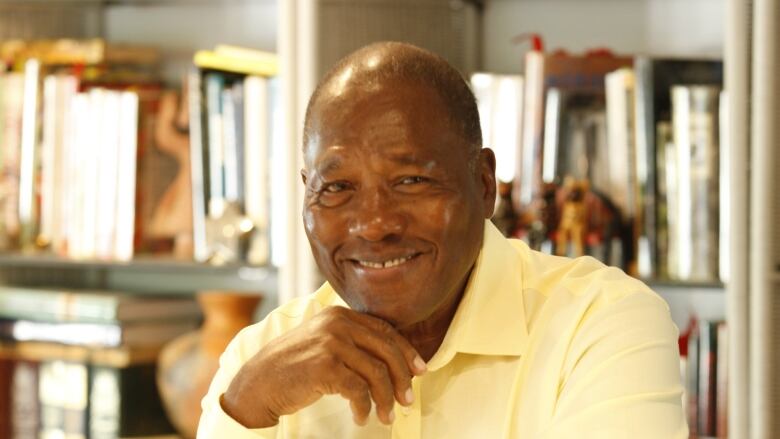 A man sits at a library table with a wry grin. Stacks of books can be seen behind him.