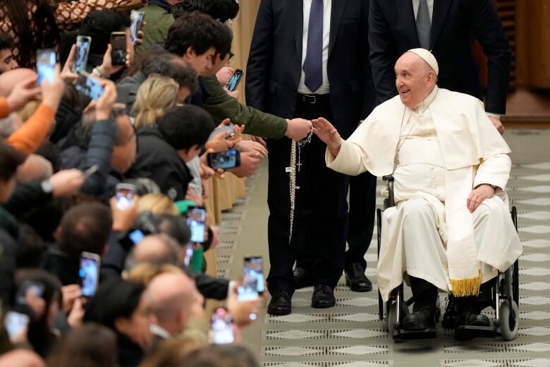 Pope Francis, in a wheelchair, shakes hands with a man in a crowd at the Vatican.