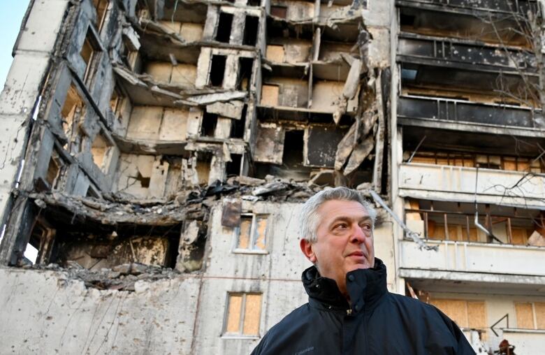 A man is shown in medium closeup in front of a damaged urban residential building.