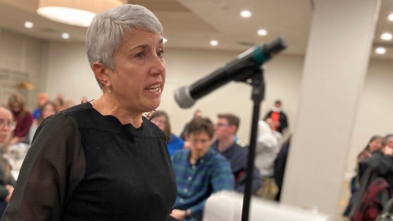 A woman with short white hair speaks into a microphone, crowd in the background