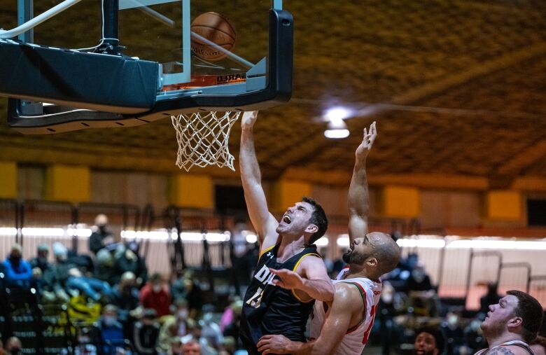 A basketball player in a dark uniform jumps for a rebound in front of a player in a white uniform.