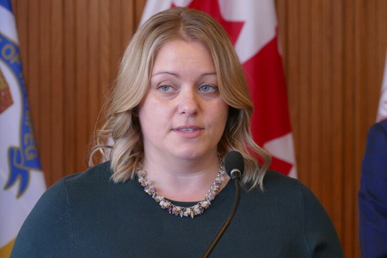 A woman speaks into a microphone in a close-up shot. Behind her there are several flags.