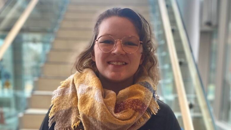 A woman in a yellow scarf smiles into the camera, with stairs in the background.