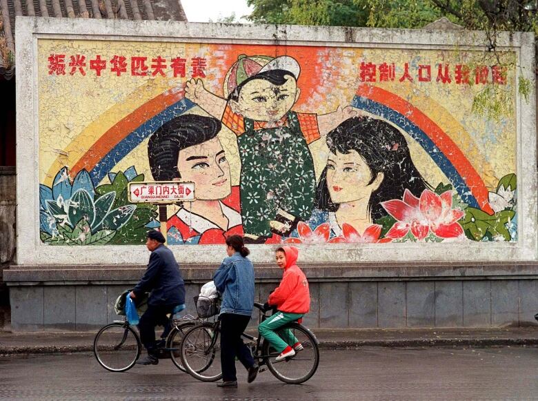 Three people on bicycles ride past a decaying mural showing a man, woman and one child. 